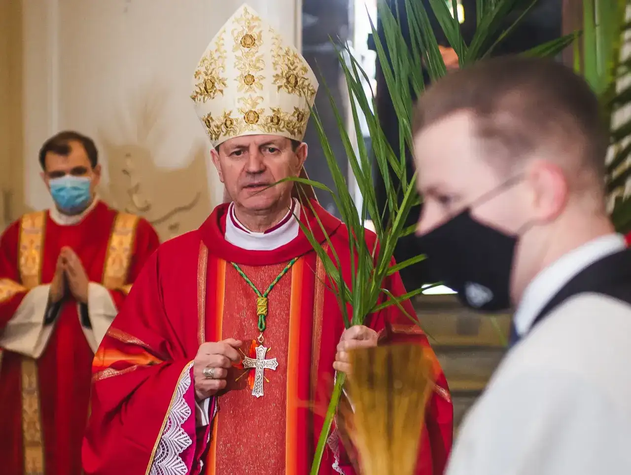 Biskup w Polsce, w czerwonej szacie i mitrze, trzyma gałązki palmowe. Obok klęczy wierny.