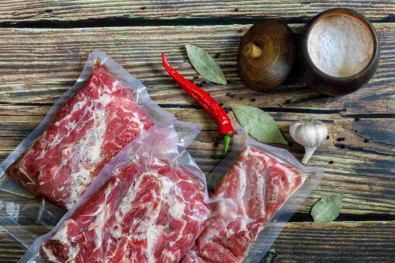 Vakuumverpacktes Rindfleisch, das sauer riecht, liegt neben einem roten Chili, Knoblauch und Lorbeerbl&auml;ttern auf einem Holztisch.