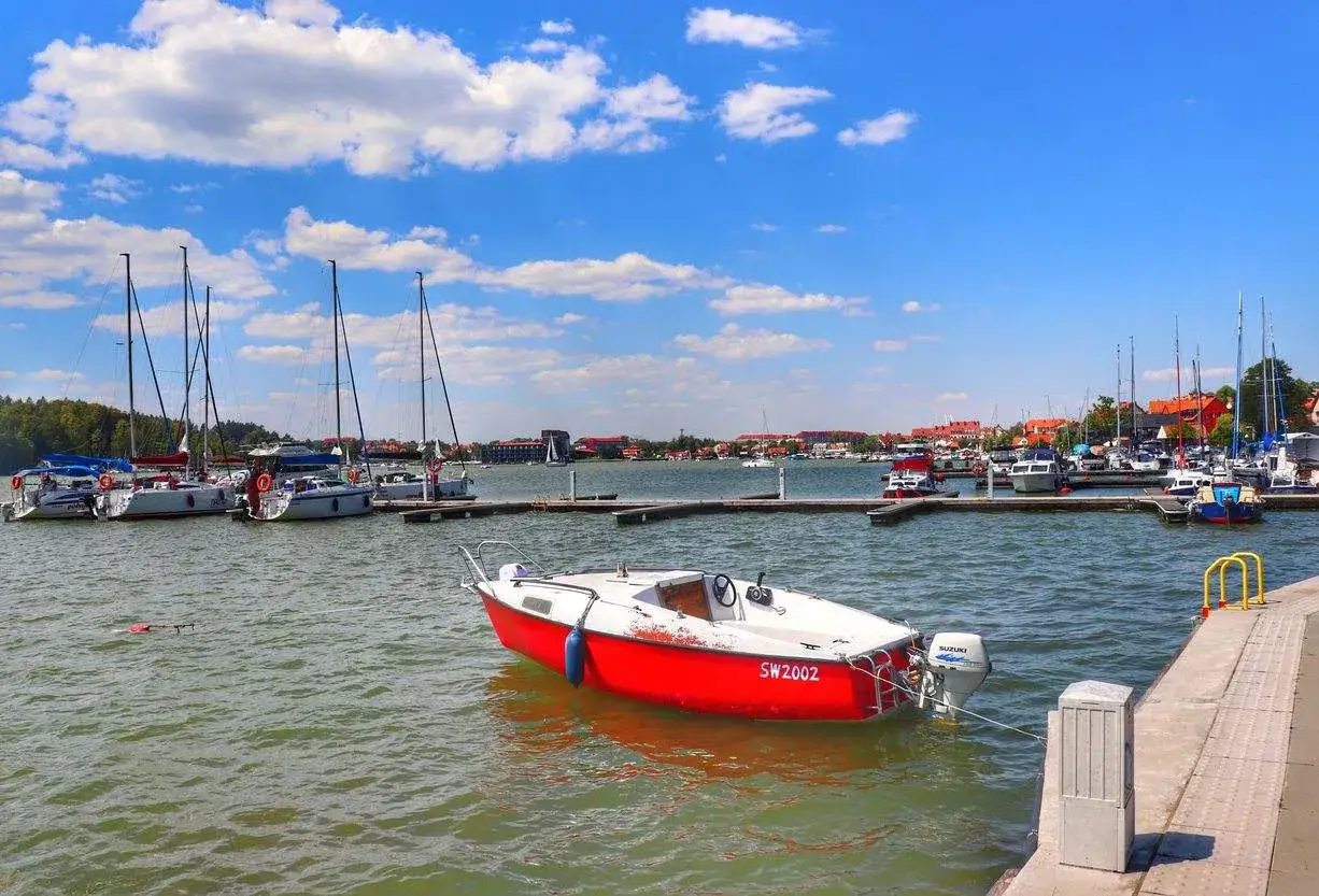 Czerwona ł&oacute;dź kołysze się na wodzie, a w tle port pełen żagl&oacute;wek. Idealne miejsce na mikołajki atrakcje i wodne szaleństwo.