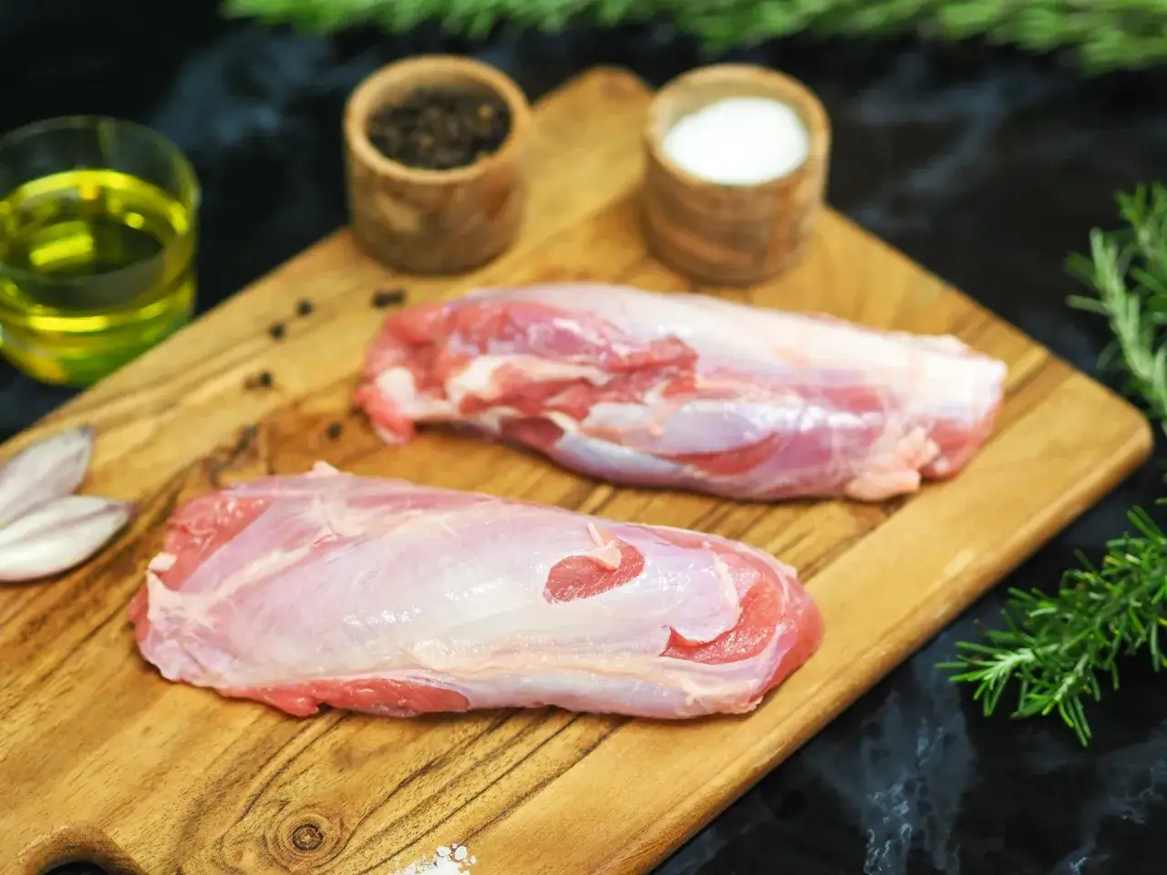 Deux morceaux de jaret crus sur une planche en bois, accompagnés d'huile d'olive, de poivre, de sel et de romarin.