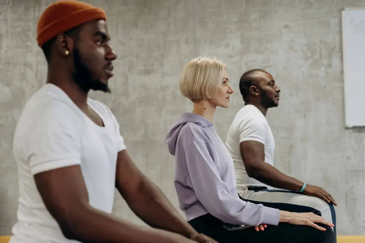 Tres personas sentadas en fila, practicando meditación. El hombre de la izquierda lleva un gorro naranja. El significado de compasión se refleja en su calma.