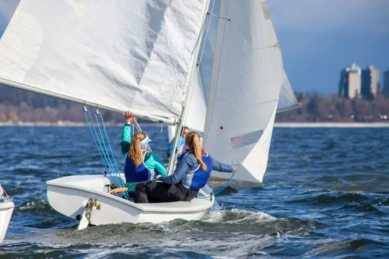 Deux jeunes femmes naviguent sur un petit type de bateau à voile, leurs voiles blanches gonflées par le vent sur une eau bleue.