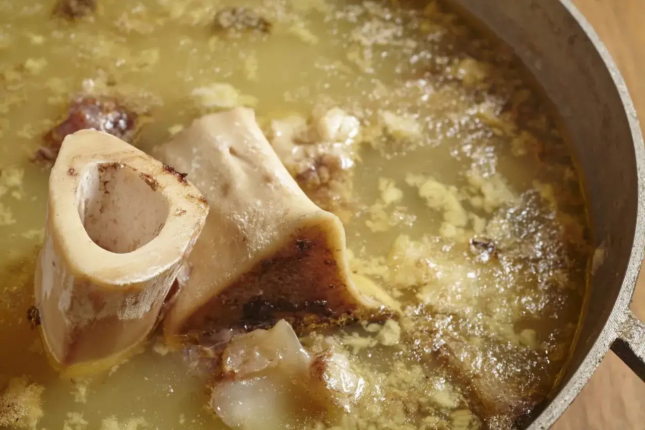 Un primer plano de un caldo de huesos para perros en una olla. Los huesos grandes y cortados est&aacute;n sumergidos en un l&iacute;quido dorado y turbio.