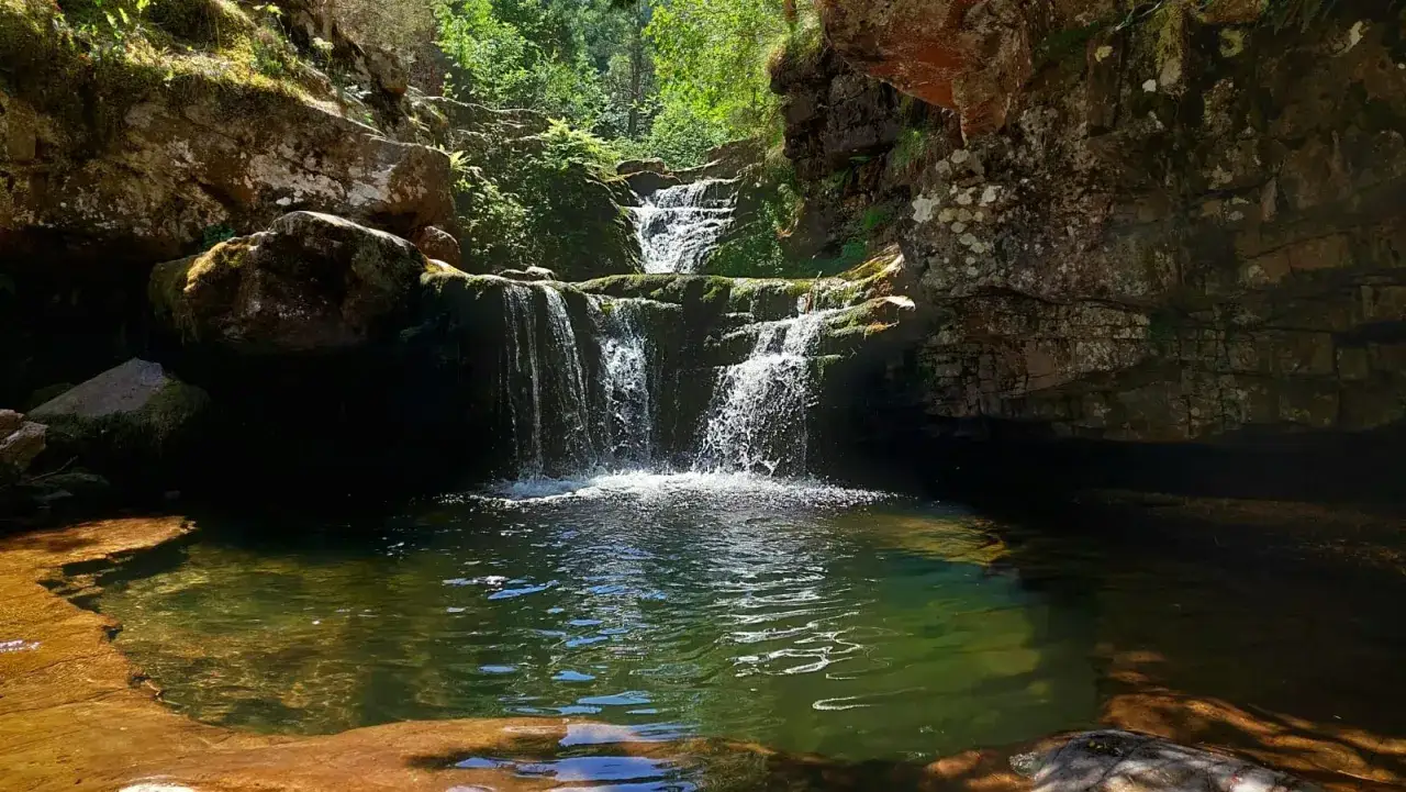 Cascada en un bosque frondoso, con rocas cubiertas de musgo y un estanque de aguas cristalinas. El **puente ra** natural de rocas permite cruzar el arroyo.
