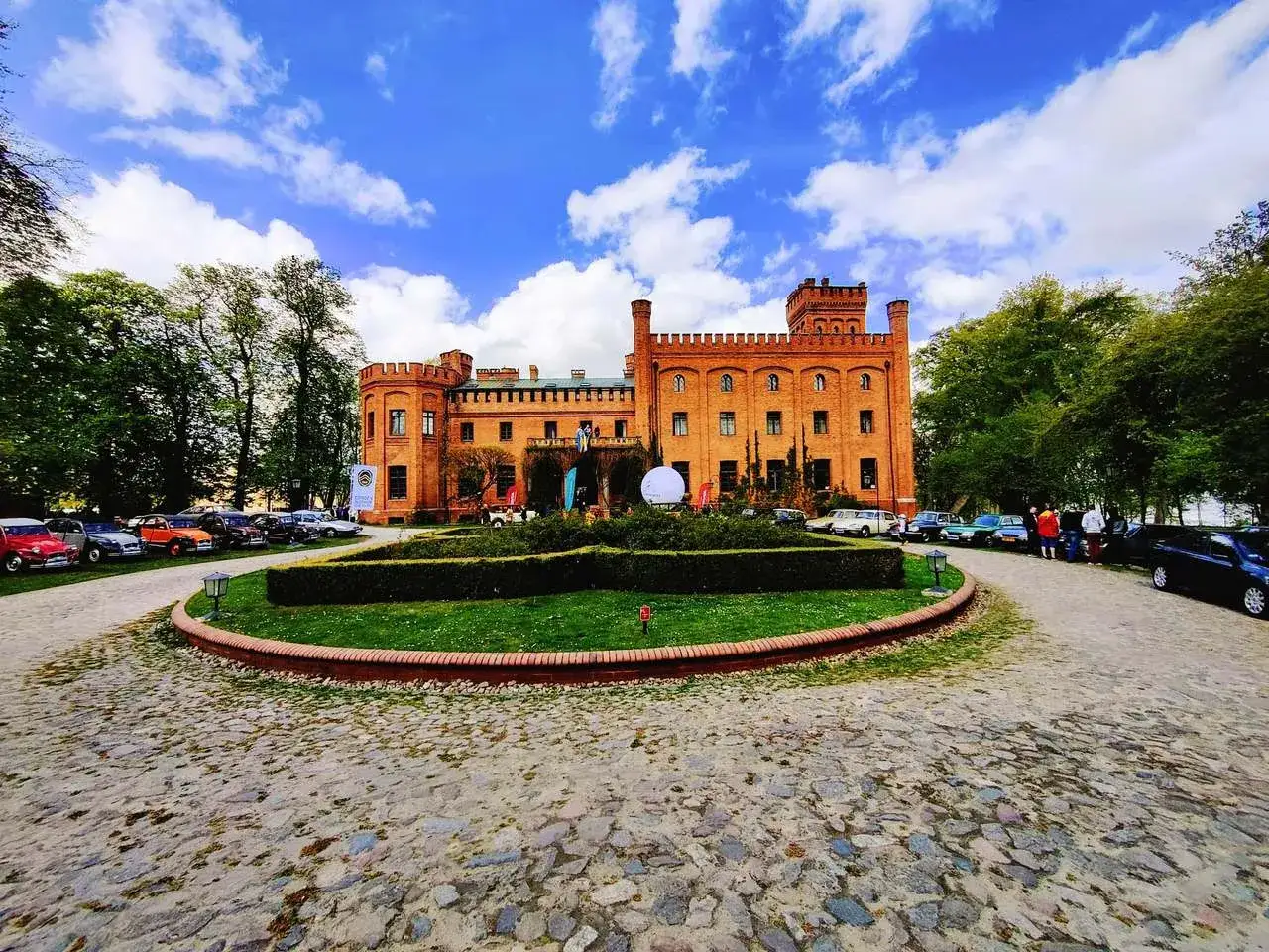 Zamek w Rzucewie, ceglana budowla z wieżyczkami, otoczony zielenią i brukowanym podjazdem, przy kt&oacute;rym stoją zabytkowe samochody.