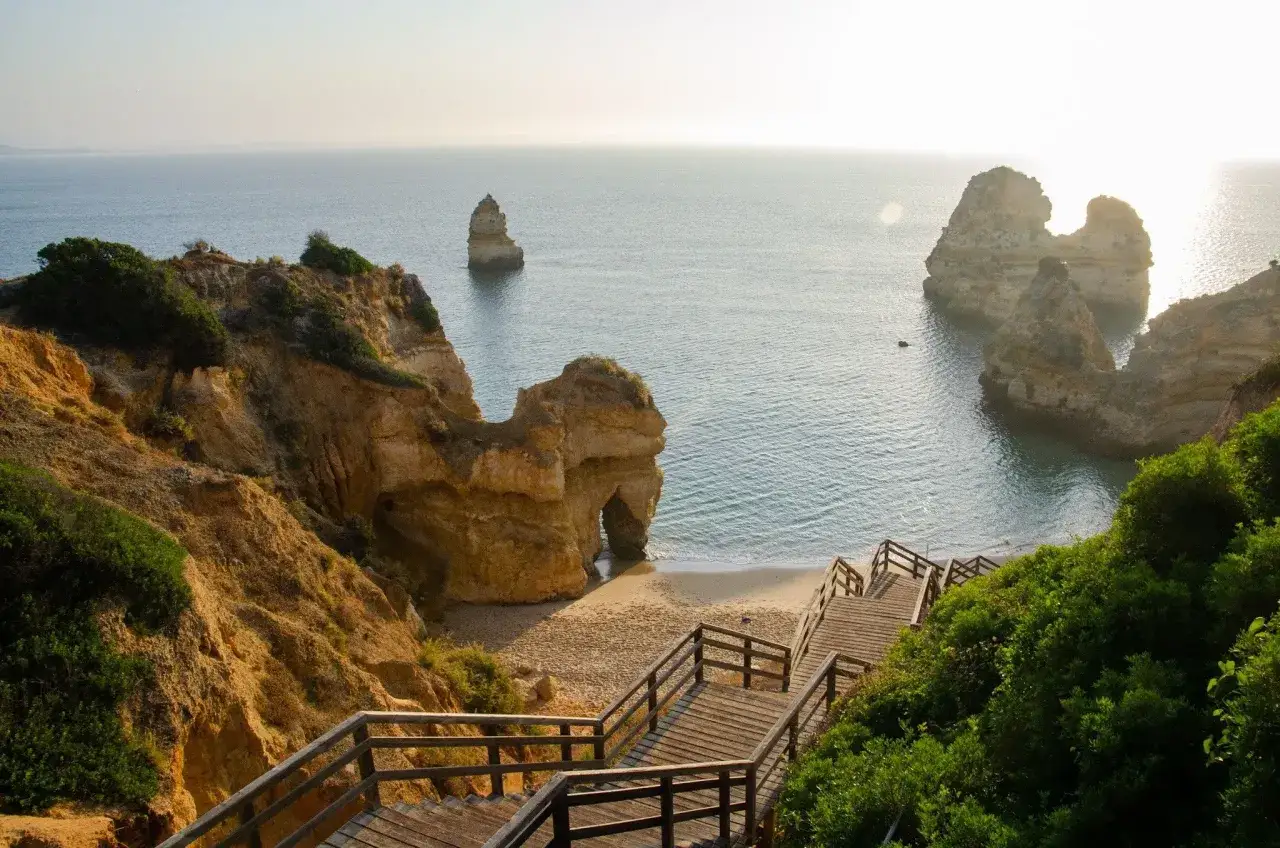 Sonnige Felsformationen und eine Holztreppe f&uuml;hren zu einem Strand an der portugiesischen K&uuml;ste. Perfektes Wetter in Portugal im Oktober.
