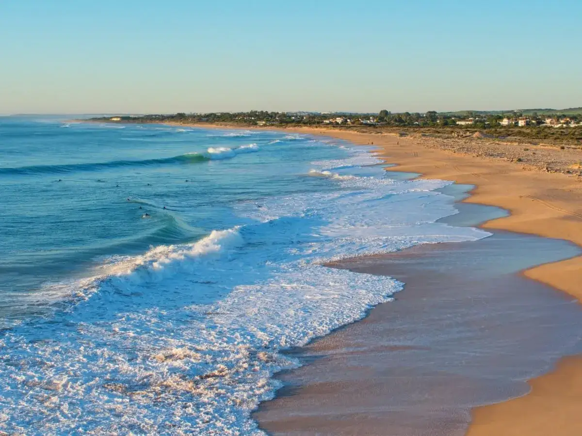 Der conil de la frontera strand mit sanften Wellen und Surfern im t&uuml;rkisfarbenen Meer. Im Hintergrund eine K&uuml;stenlinie mit H&auml;usern.