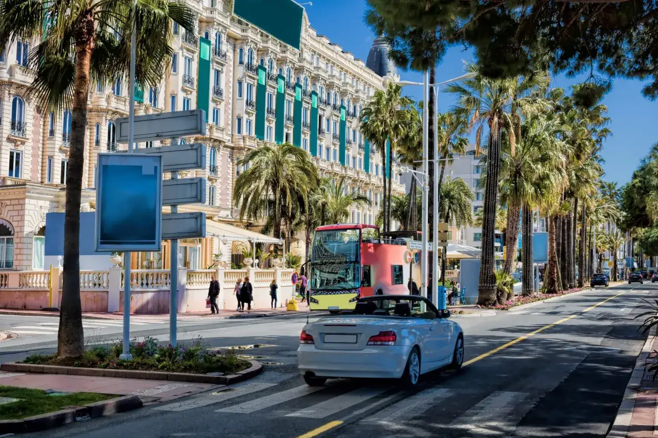 Biały kabriolet na pasach, obok luksusowy hotel i palmy. Wygląda jak droga dojazdowa do Cannes lotnisko.