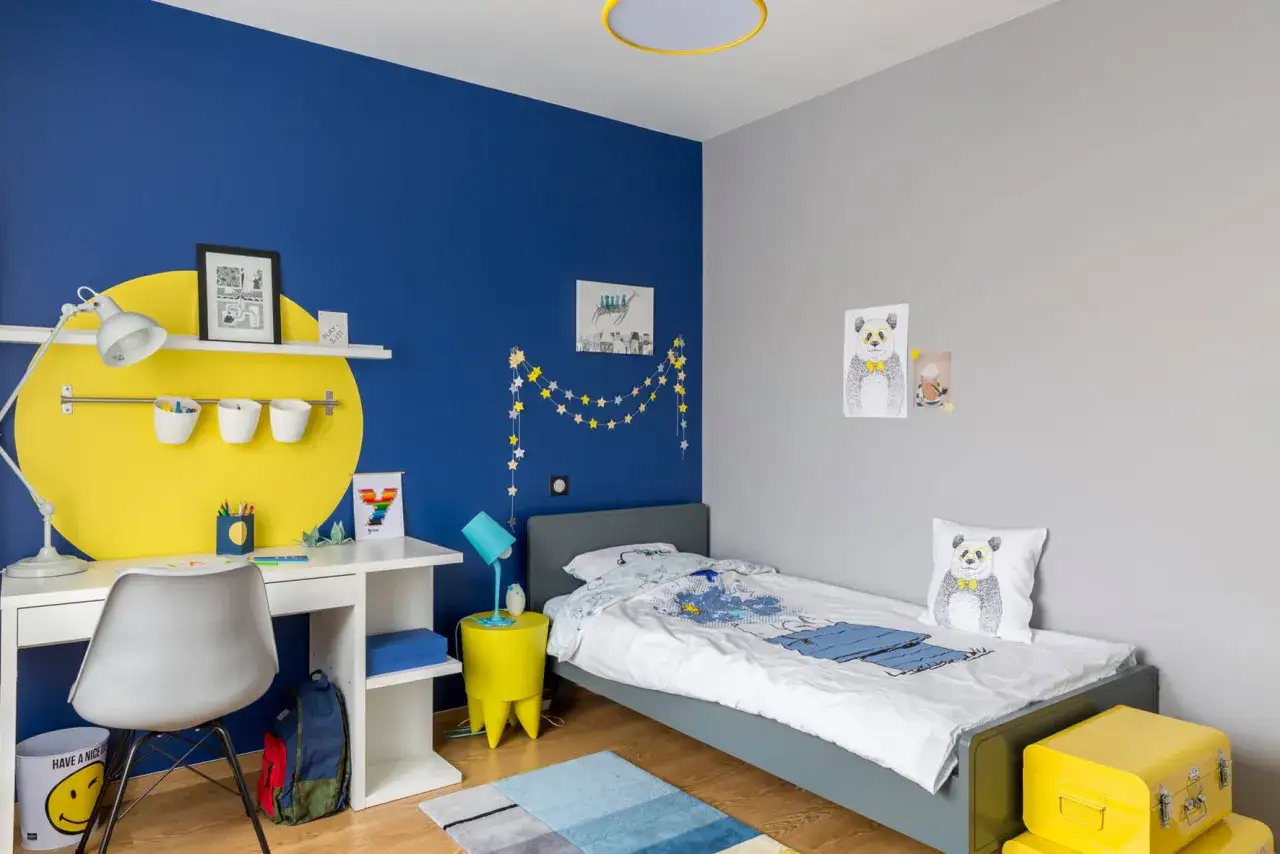 Cameretta con parete blu vivace e decorazioni a stelle. Scrivania bianca, letto grigio e dettagli gialli per idee pittura cameretta.