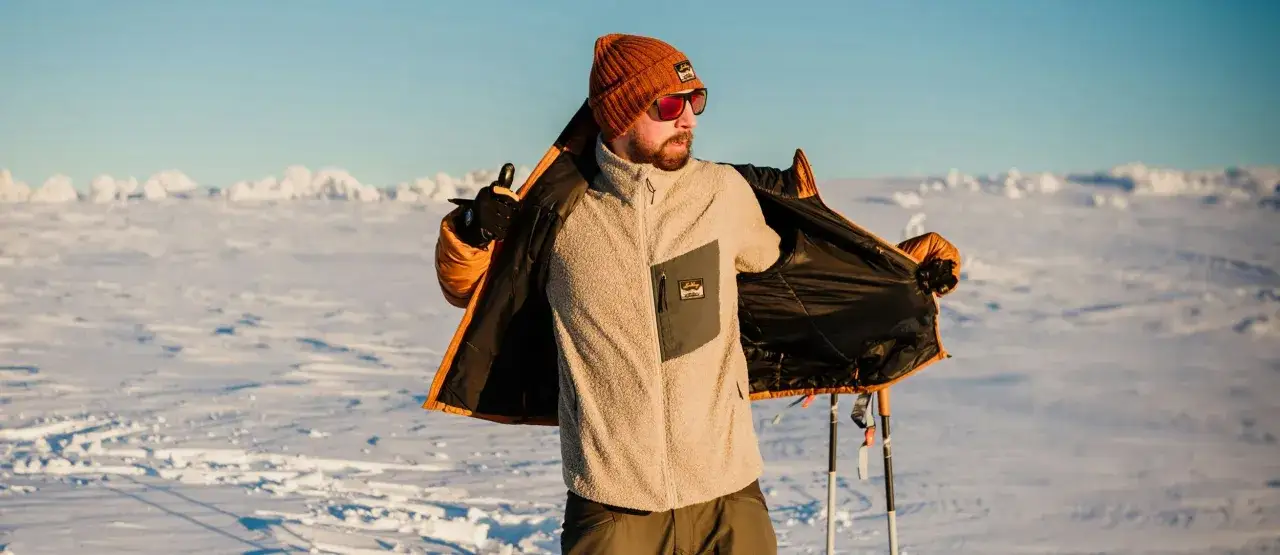 Man i orange m&ouml;ssa och solglas&ouml;gon &ouml;ppnar en jacka som visar att han har lager p&aring; lager. Skidstavar syns i bakgrunden i ett sn&ouml;igt landskap.