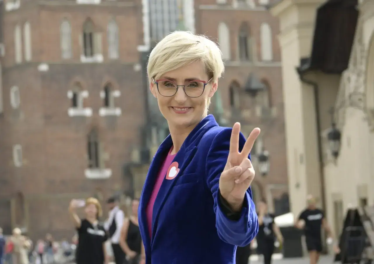 Katarzyna Matusik w lipcu pozdrawia znakiem pokoju, ubrana w niebieski żakiet z sercem na klapie.