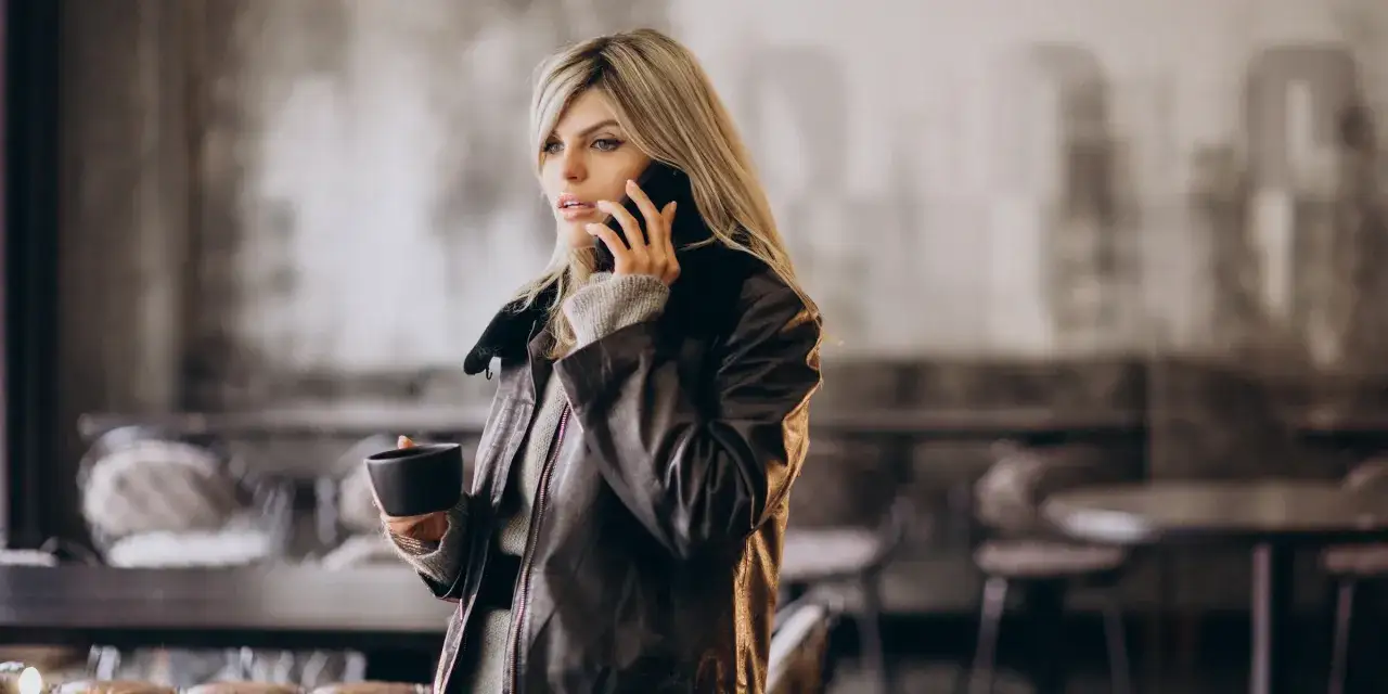 Femme blonde en veste en cuir, tenant une tasse et parlant au t&eacute;l&eacute;phone dans un caf&eacute;.