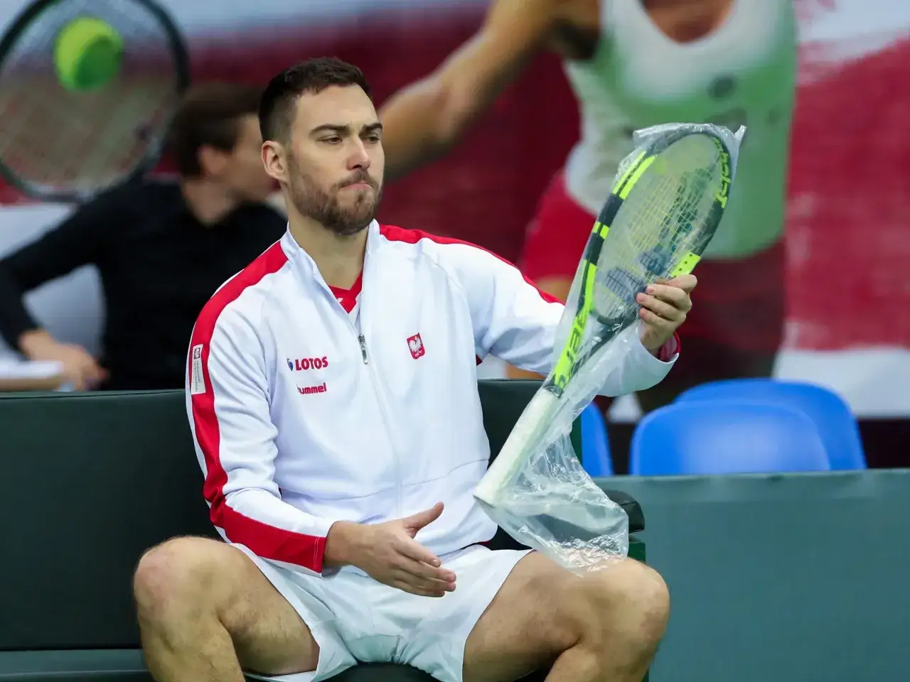 Jerzy Janowicz, w białej bluzie z polskim orłem, siedzi na ławce, trzymając rakietę tenisową w folii.