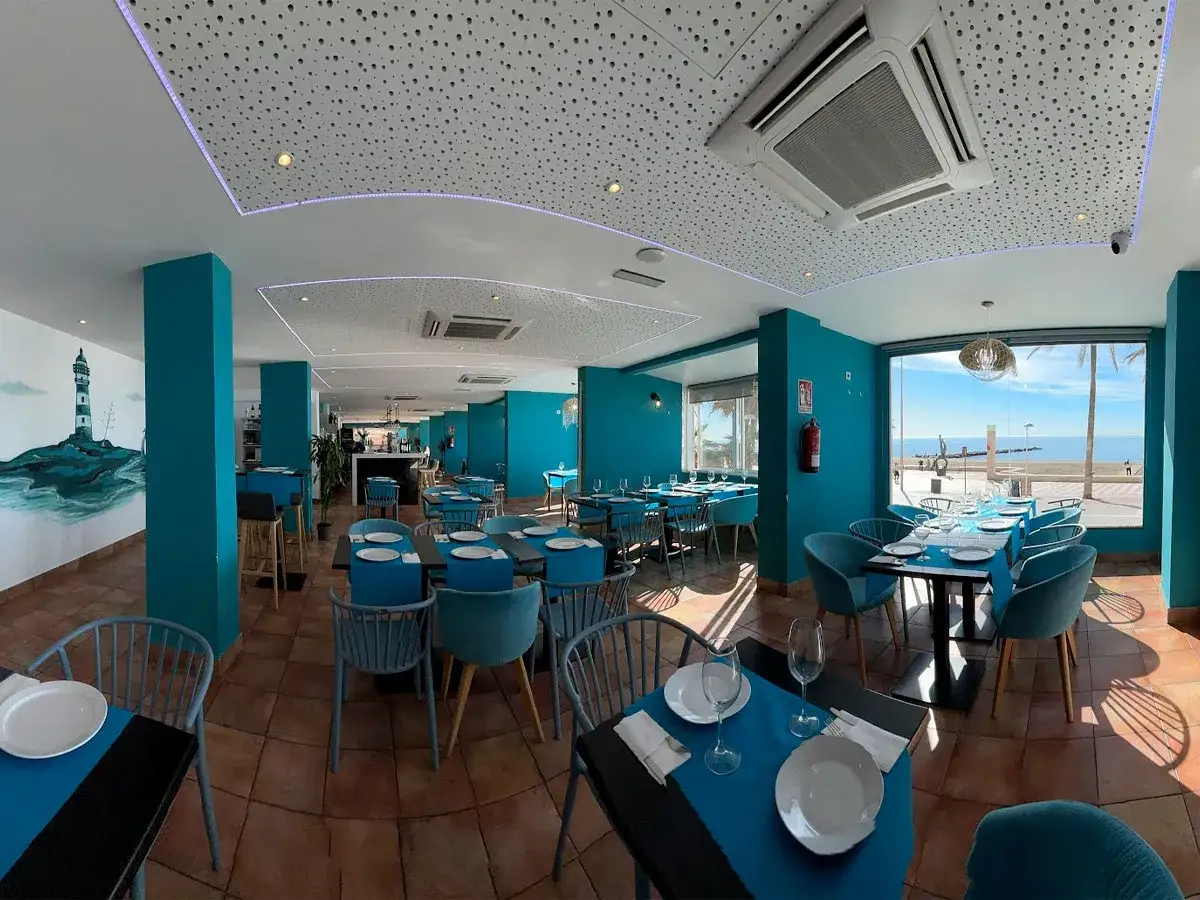 Interior del chiringuito La Sal, con mesas y sillas azules, mural de faro y vistas al mar.