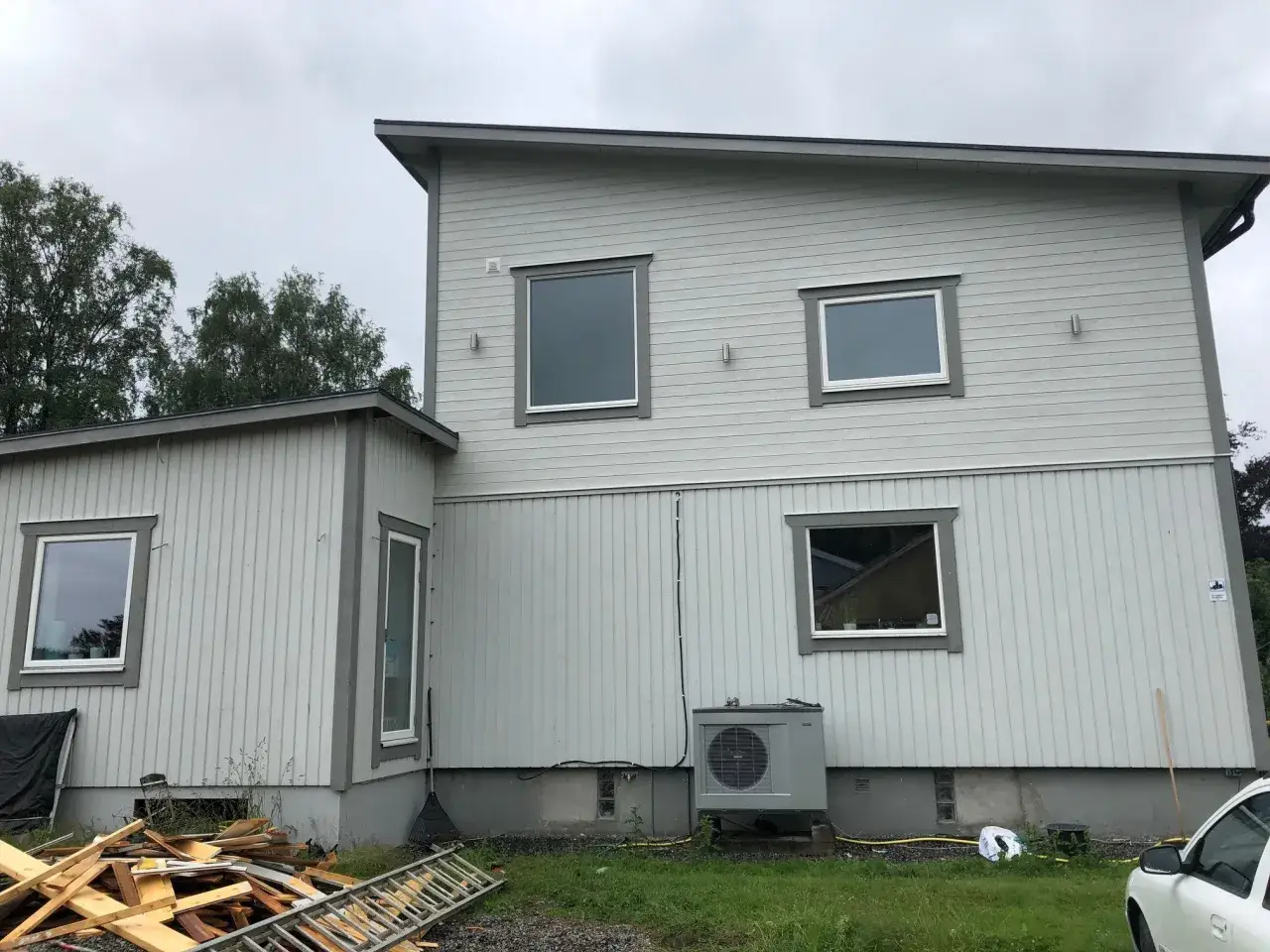 Grått hus med vita fönster och en luftvärmepump. Byggmaterial och en stege ligger på marken, kanske förberedelser för taklyft pris.