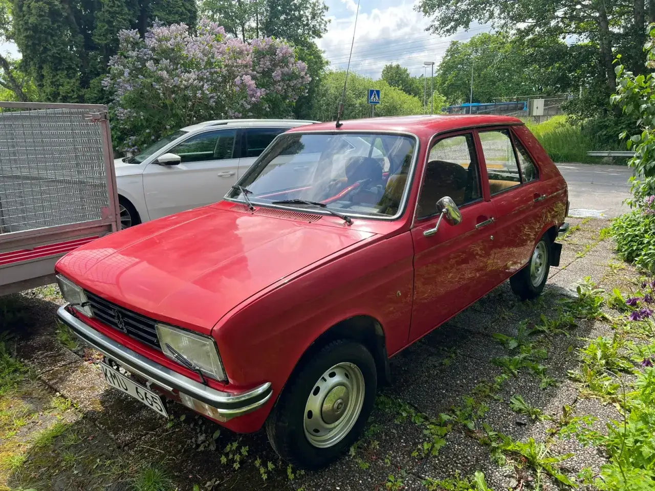 Röd Peugeot 104 parkerad utomhus, med en vit kombi och en trailer bredvid. Blommor och träd i bakgrunden.