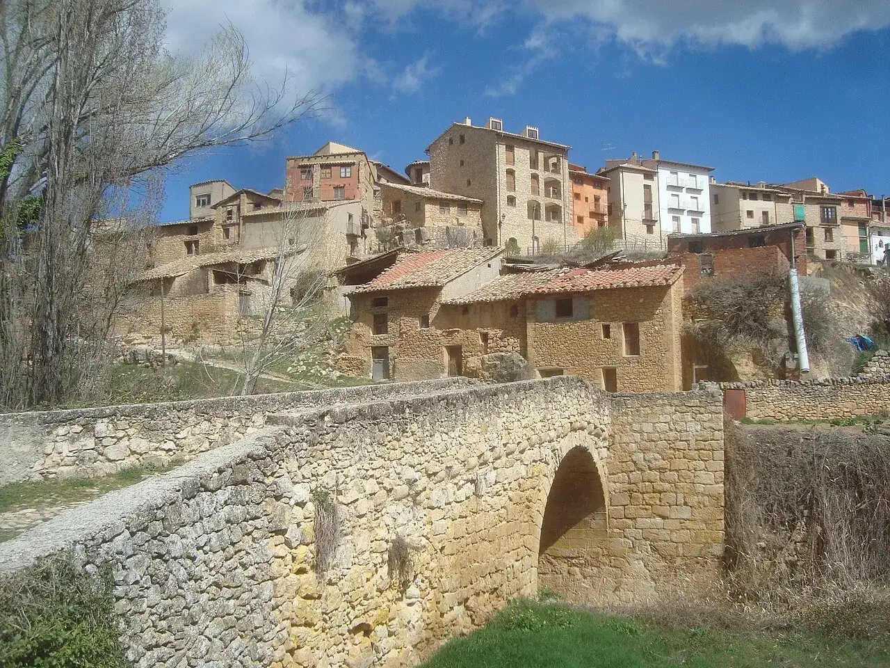 Un puente de piedra cruza un arroyo, llevando a un pueblo con casas de piedra y tejados rojos. Al fondo, se alza la **torre de las arcas**, vigilante del paisaje.