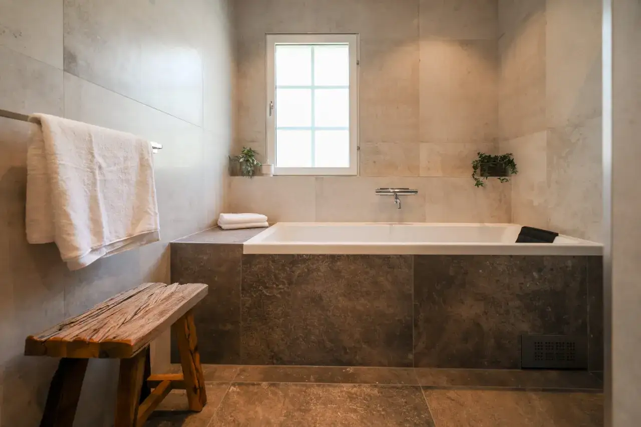Ett badrum med spa-känsla. Ljusgrå kakel på väggarna, mörkare stenliknande kakel runt badkaret. En träpall och en handduk bidrar till spa inredning.