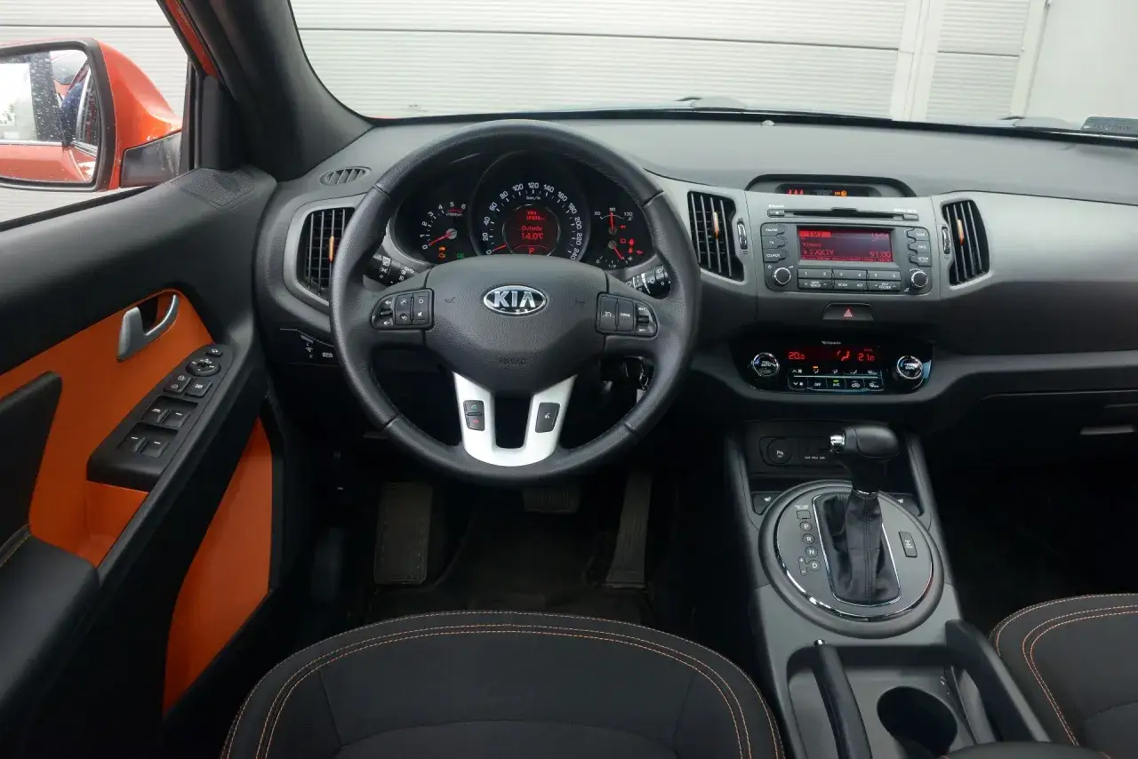 Wnętrze Kia Sportage 1.7 CRDi z pomarańczowymi akcentami. Widoczny kokpit, kierownica, radio i automatyczna skrzynia biegów.