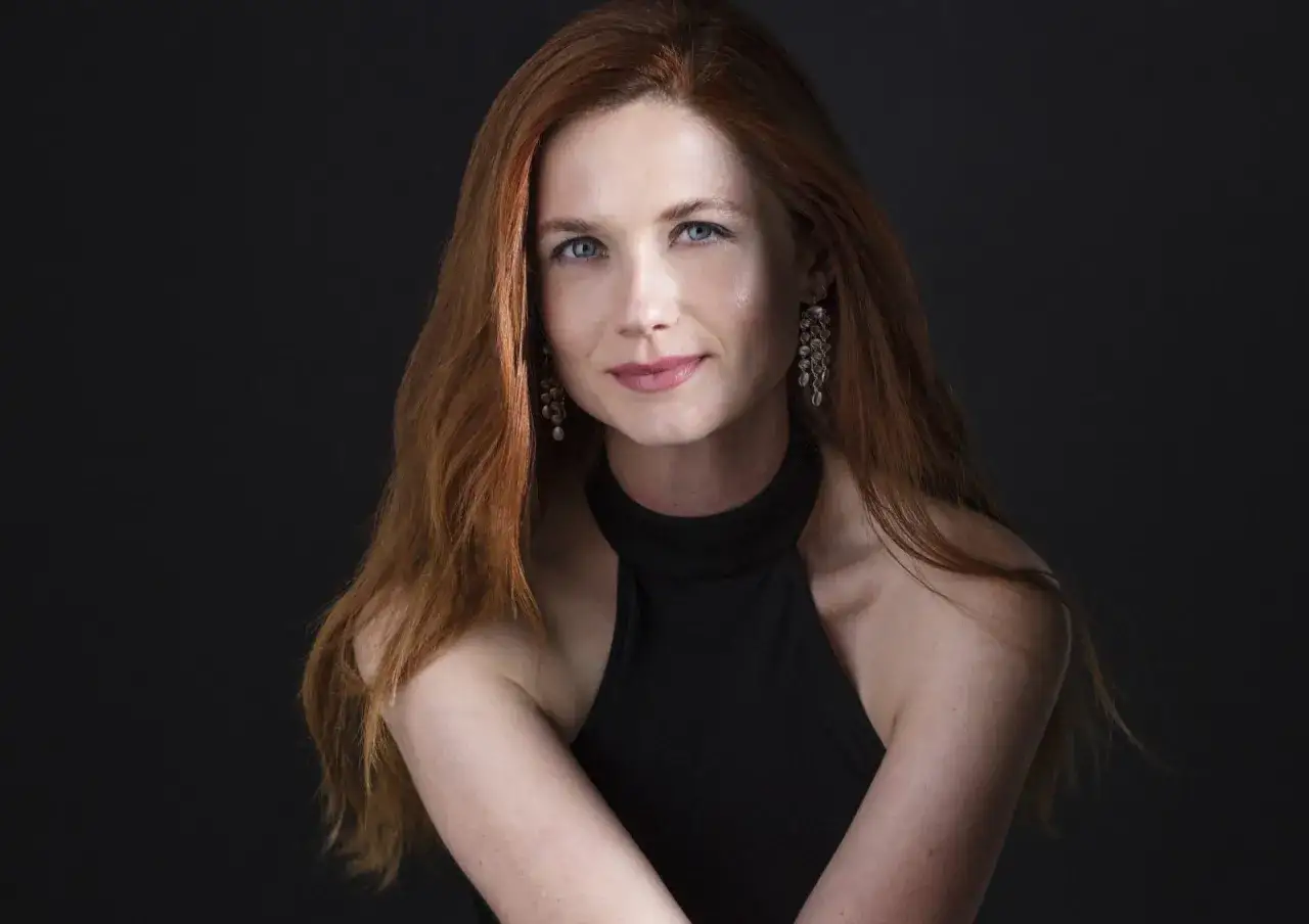Bonnie Wright wiek: rudowłosa kobieta z niebieskimi oczami, w czarnej sukience i wiszącymi kolczykami, na ciemnym tle.