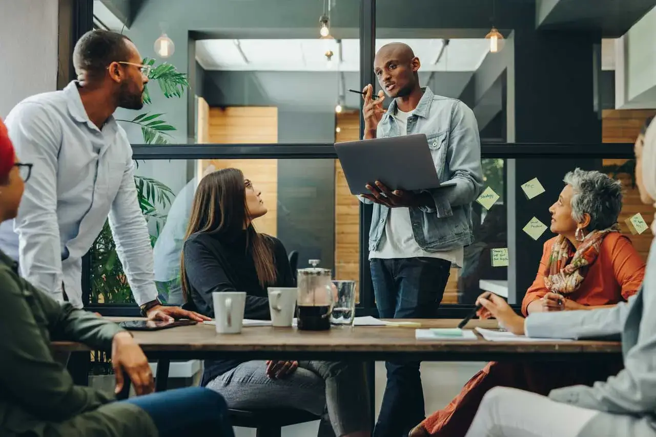 Une &eacute;quipe discute autour d'une table, un homme pr&eacute;sente un projet sur son ordinateur. L'ergonomie organisationnelle au c&oelig;ur de leur collaboration.