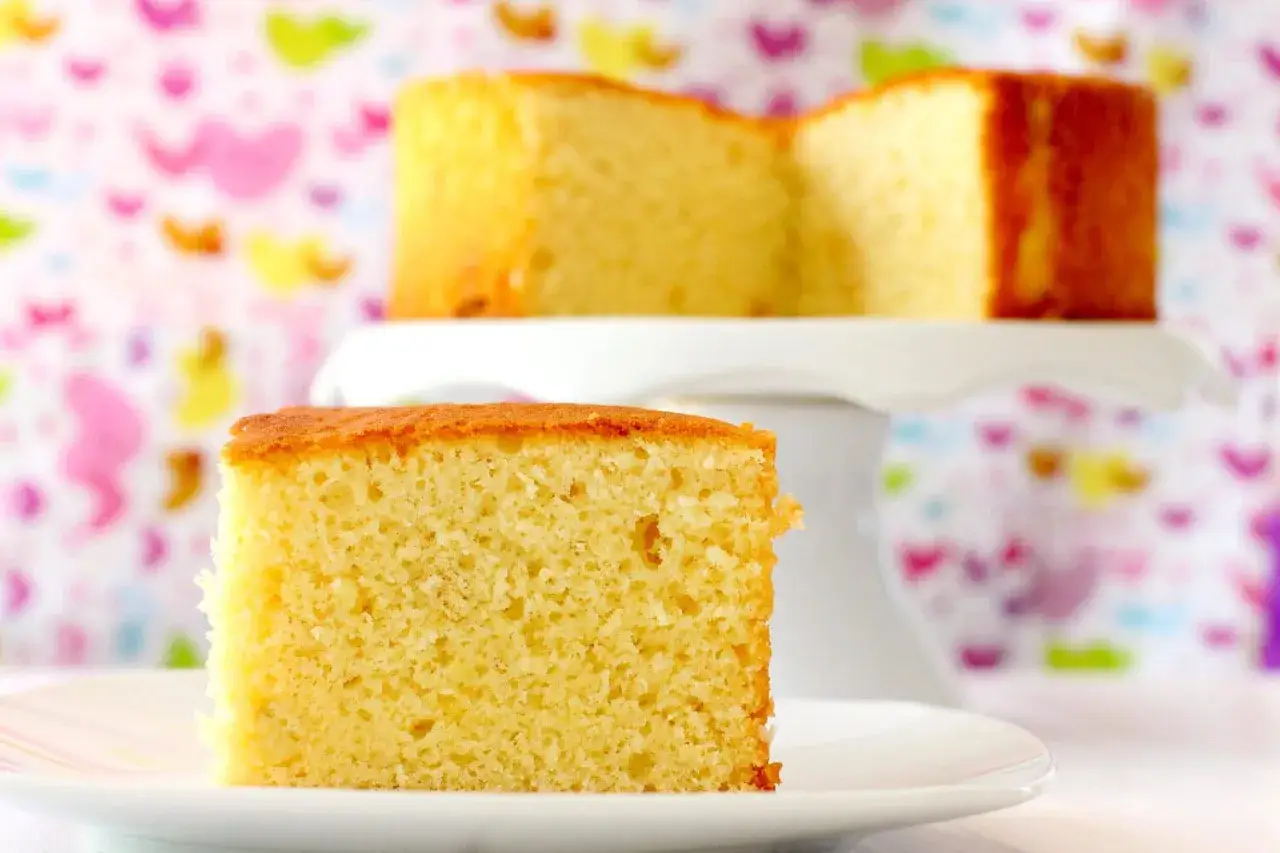 Un delicioso bizcocho sin huevo, esponjoso y dorado, servido en un plato blanco. Otro trozo se ve en un soporte para pasteles al fondo.