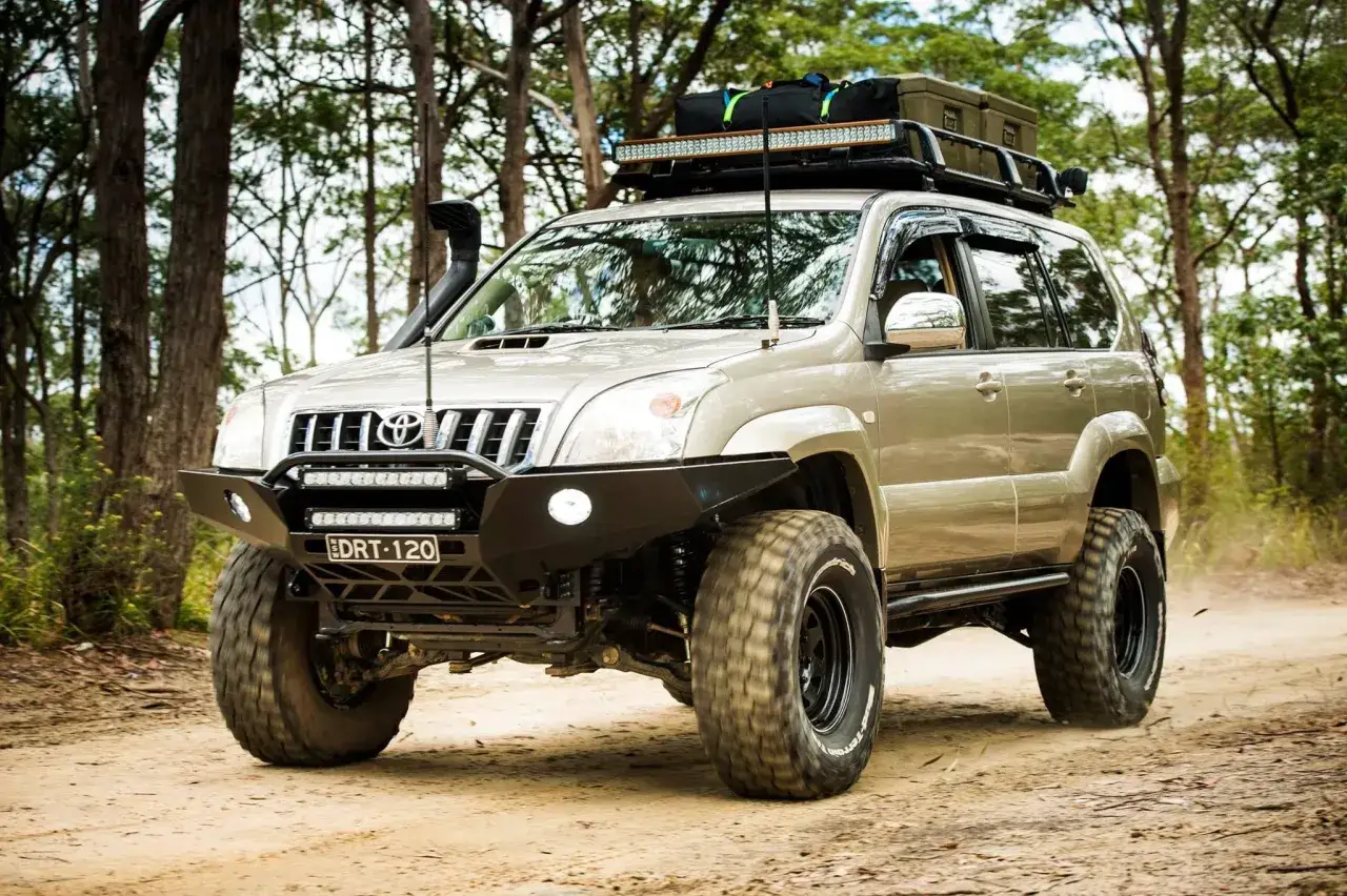 Zmodyfikowany Toyota Land Cruiser 120 z terenowym zderzakiem, bagażnikiem dachowym i snorkelami gotowy na przygodę w lesie.