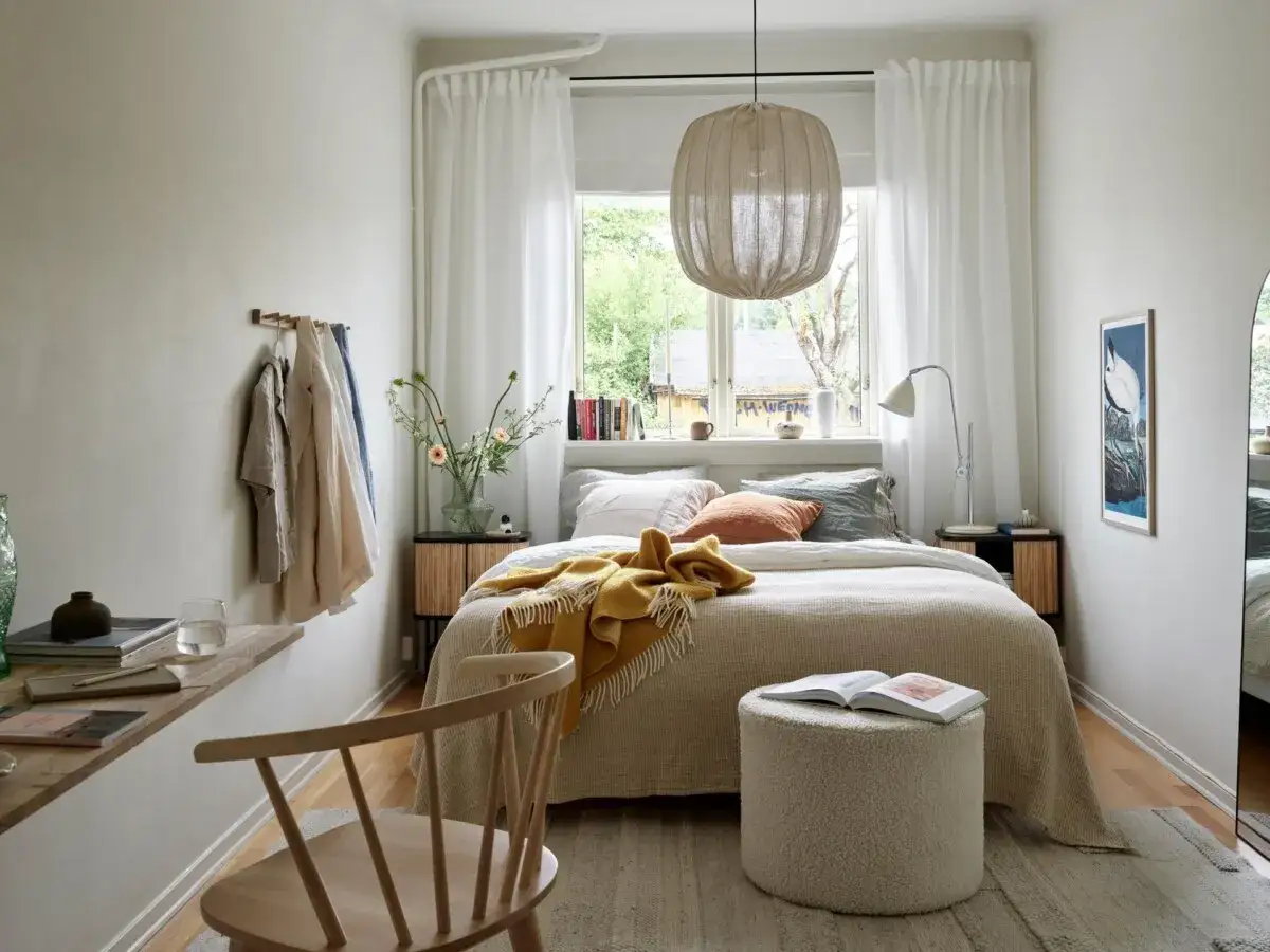Cama bajo ventana, ideas para un dormitorio acogedor. Ropa colgada, manta amarilla y un puf mullido invitan al descanso.