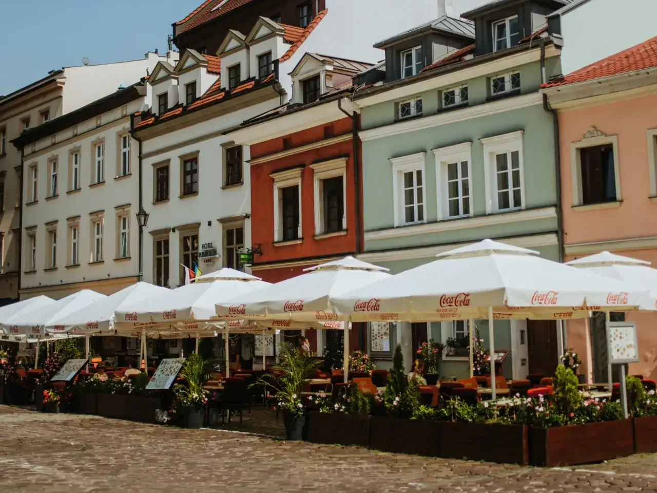 Krak&oacute;w stare miasto restauracje z białymi parasolami Coca-Cola. Kolorowe kamienice tworzą tło dla letniego ogr&oacute;dka.