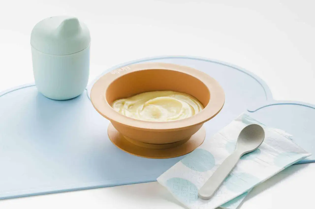 Babybrei in einem orangefarbenen Beikost-Schüsselchen, daneben ein Becher und ein Löffel auf einer blauen Unterlage.