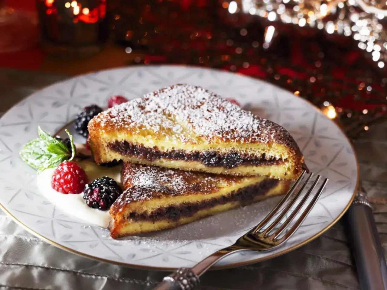 Zwei St&uuml;cke gef&uuml;llter French Toast mit Puderzucker, Beeren und Minze auf einem Teller.