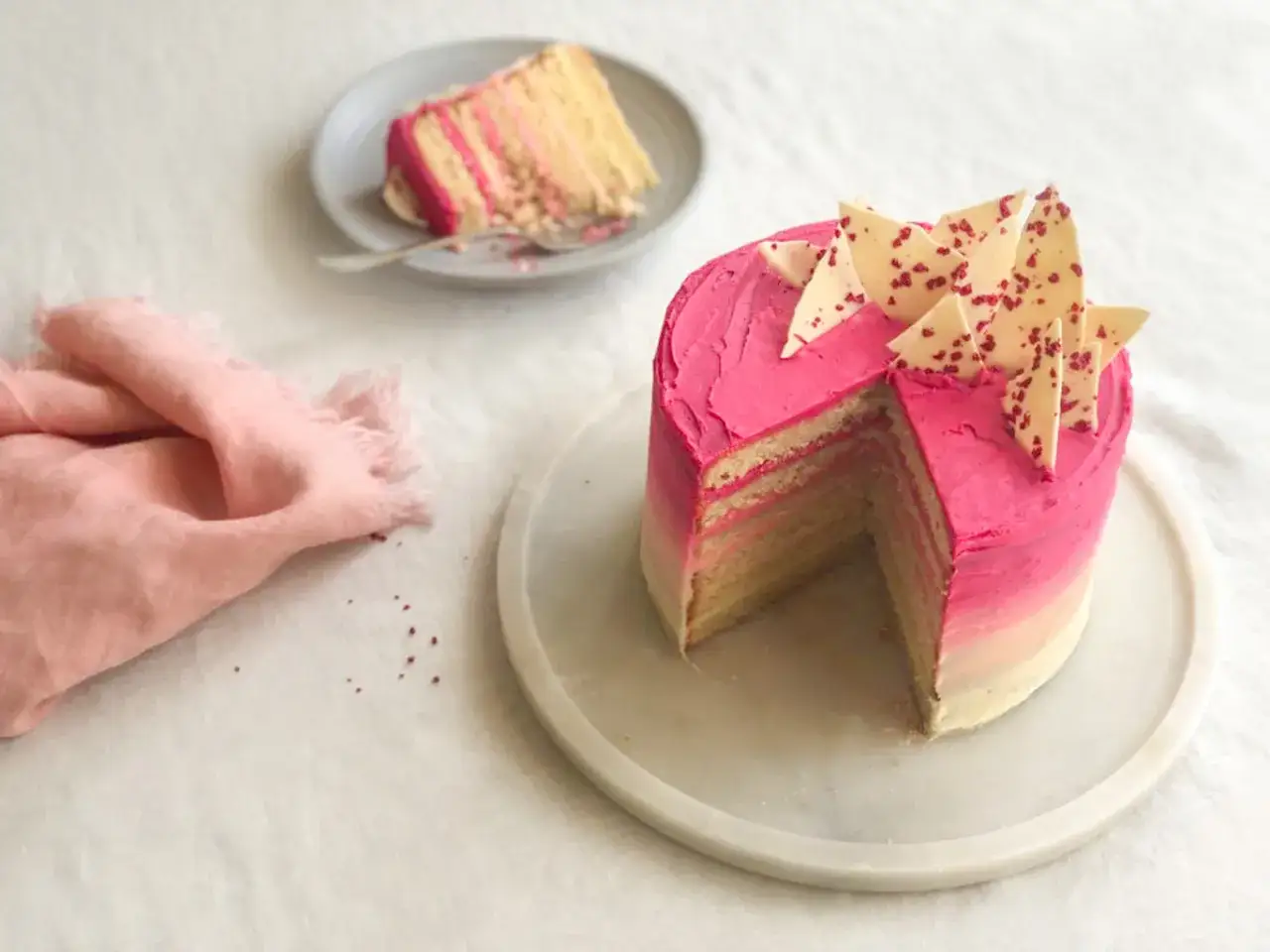 Eine mehrschichtige Buttercreme Torte mit pinkem Ombre-Frosting und weißen Schokoladenstücken. Ein Stück ist bereits auf einem Teller.