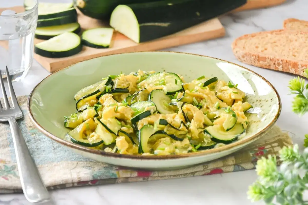 Delicioso revuelto de calabacín con huevo y cebollino, servido en un plato verde. Acompañado de pan tostado y un vaso de agua.