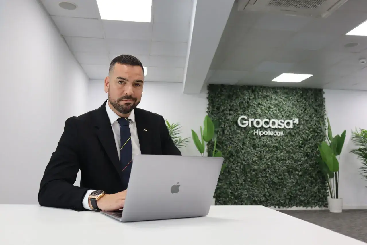 Hombre de traje trabajando en port&aacute;til. Detr&aacute;s, un muro verde con el logo de Grocasa Hipotecas. Buscando grocasa opiniones para tu hipoteca.