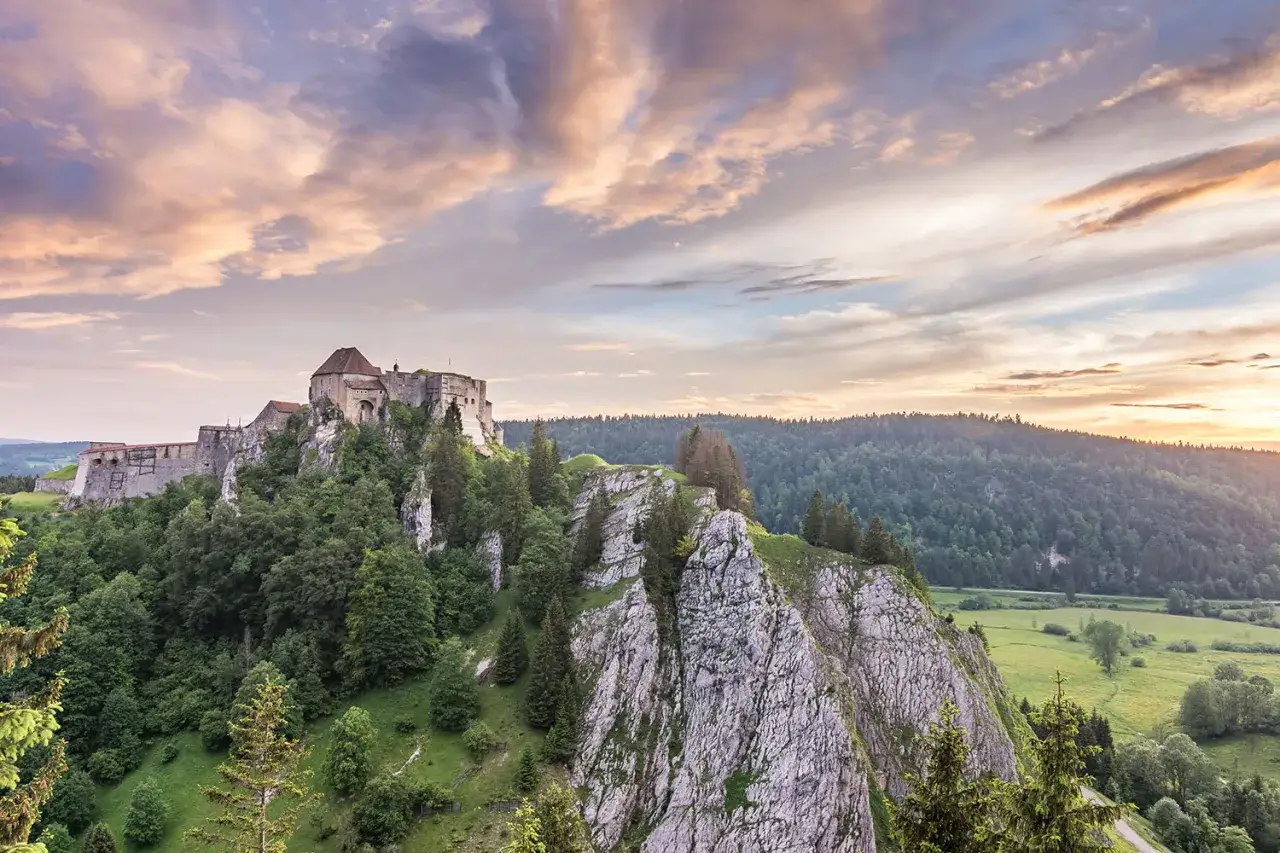 Ch&acirc;teau perch&eacute; sur un &eacute;peron rocheux, dominant un paysage verdoyant du haut Doubs au coucher du soleil. Nuages color&eacute;s dans le ciel.
