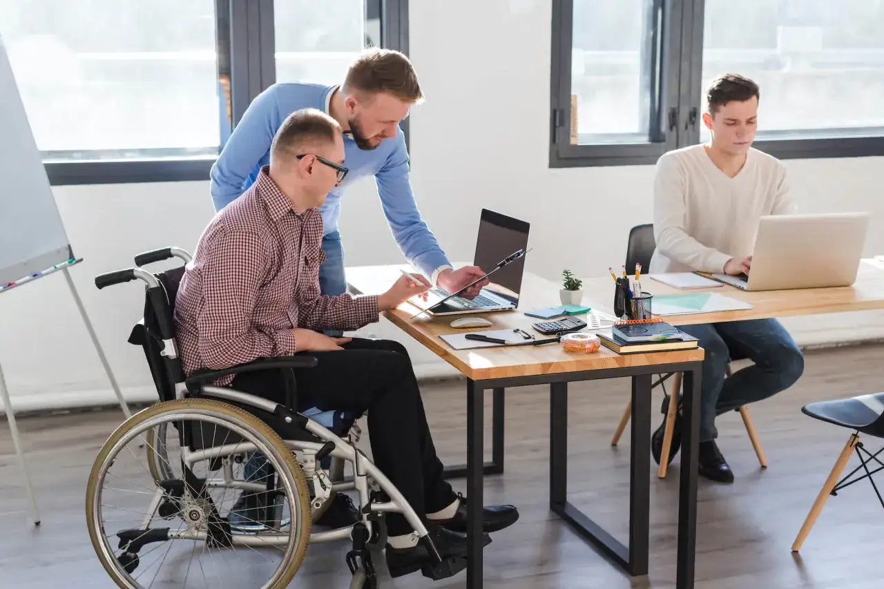 Dw&oacute;ch mężczyzn pracuje przy biurku. Jeden siedzi na w&oacute;zku inwalidzkim, drugi stoi obok. Trzeci mężczyzna pracuje przy laptopie przy innym biurku. To przykład efektywnego czasu pracy osoby niepełnosprawnej.