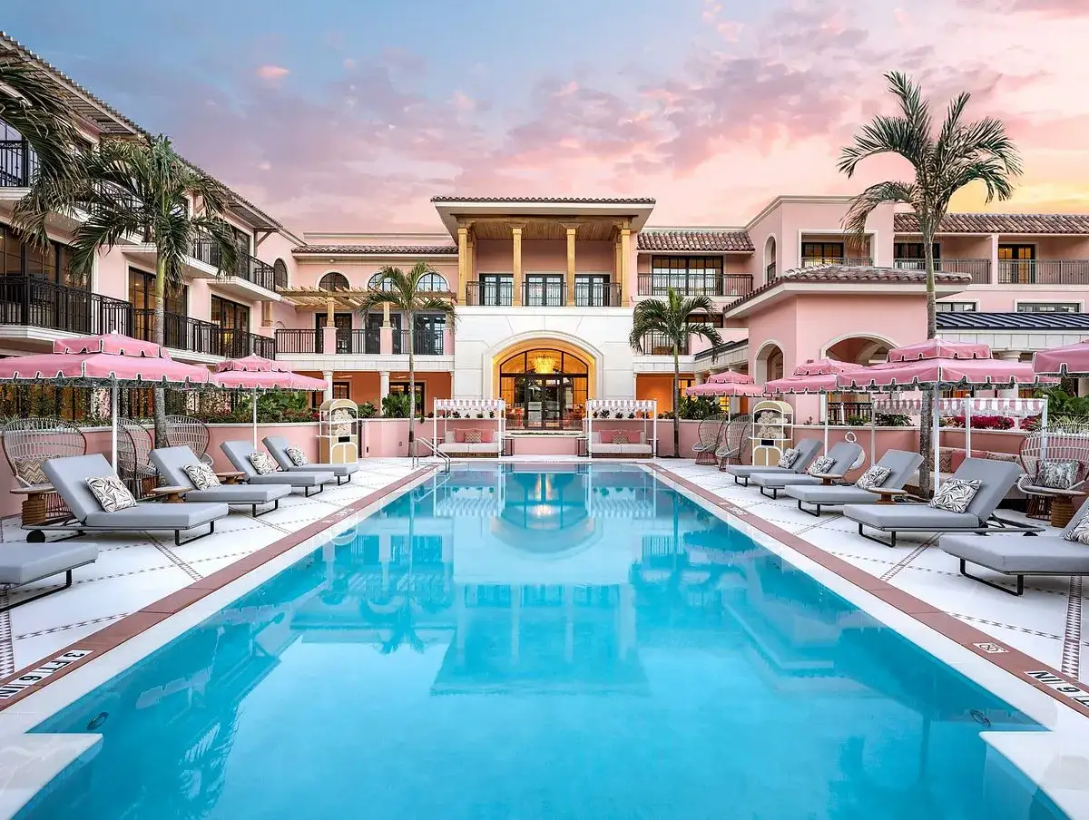 Ein luxuriöser Poolbereich mit Liegen und Sonnenschirmen vor einem rosafarbenen Gebäude. Der Himmel ist in zarten Rosa- und Orangetönen gefärbt, was an einen Sonnenuntergang erinnert. Hier könnte man sich wie ein **delphin imperial** fühlen.