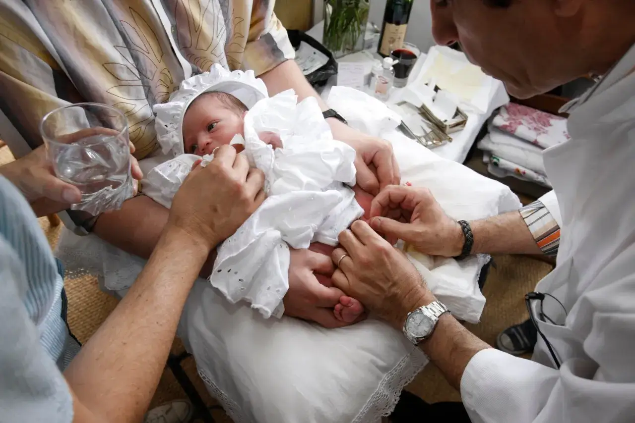 Un bébé en tenue blanche est tenu par des mains lors d'une circoncision juive. Le déroulement de ce rituel est visible avec des instruments sur la table.