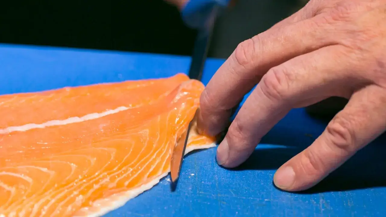 Eine Hand löst mit einem Messer die Haut vom Lachs auf einem blauen Schneidebrett.