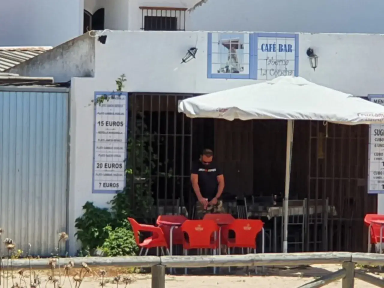 Un hombre mira su móvil frente a la Taberna La Concha, con sillas rojas y un menú de precios visibles.