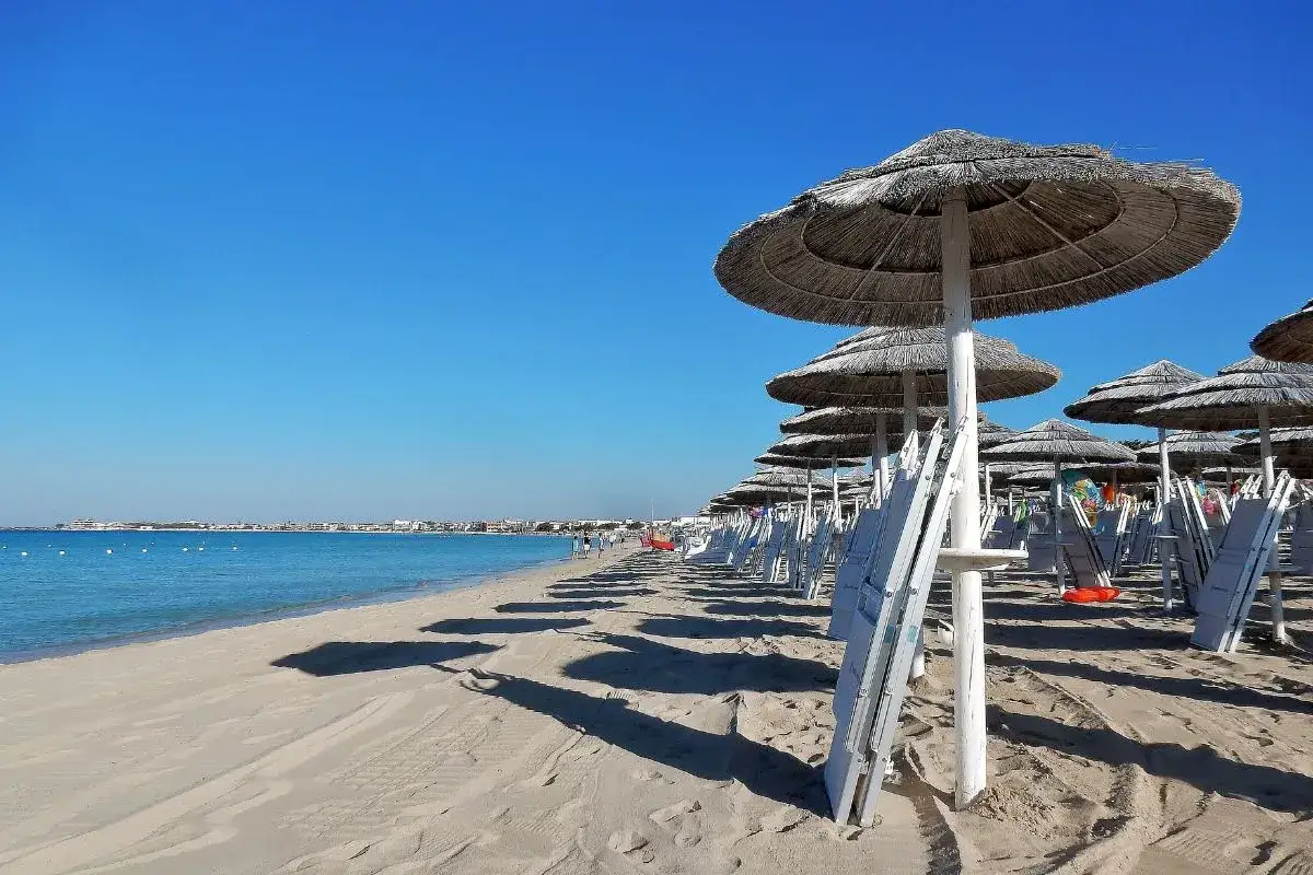 Ombrelloni di paglia e lettini bianchi su una spiaggia sabbiosa, con il mare azzurro e il cielo limpido. Il **miglior lido Torre Lapillo** offre relax e comfort.