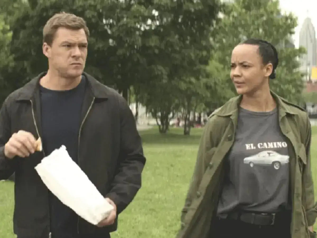 Reacher obsada: Alan Ritchson i Maria Sten idą przez park. On trzyma białą torbę, ona ma koszulkę z napisem