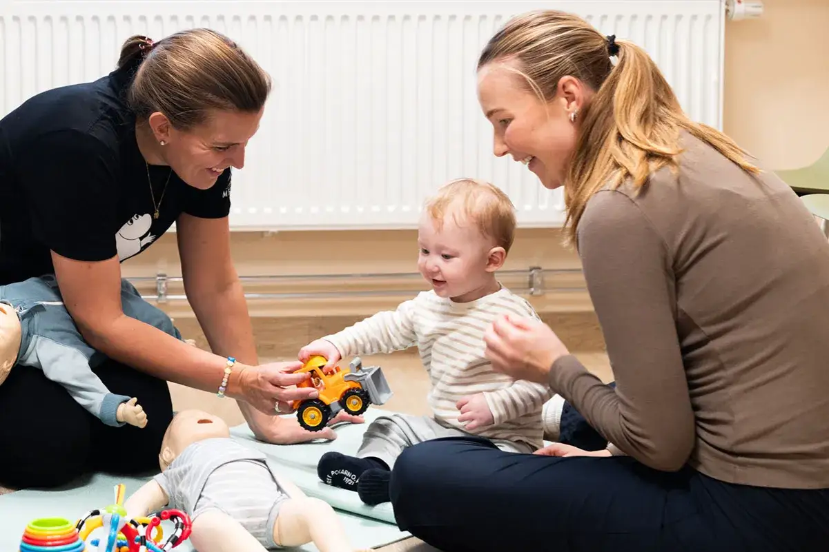 Tv&aring; kvinnor leker med en bebis och dockor. Bebisen tar en leksaksgr&auml;vmaskin, kanske en framtida ingenj&ouml;r med adhd.