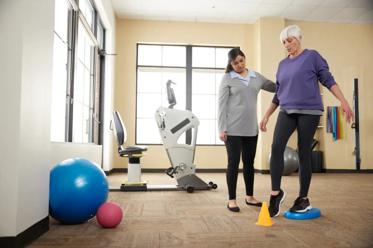 Eine &auml;ltere Frau &uuml;bt Gleichgewicht auf einem Balancekissen, w&auml;hrend eine Betreuerin hilft. Dies ist ein Beispiel f&uuml;r Senioren Sport.