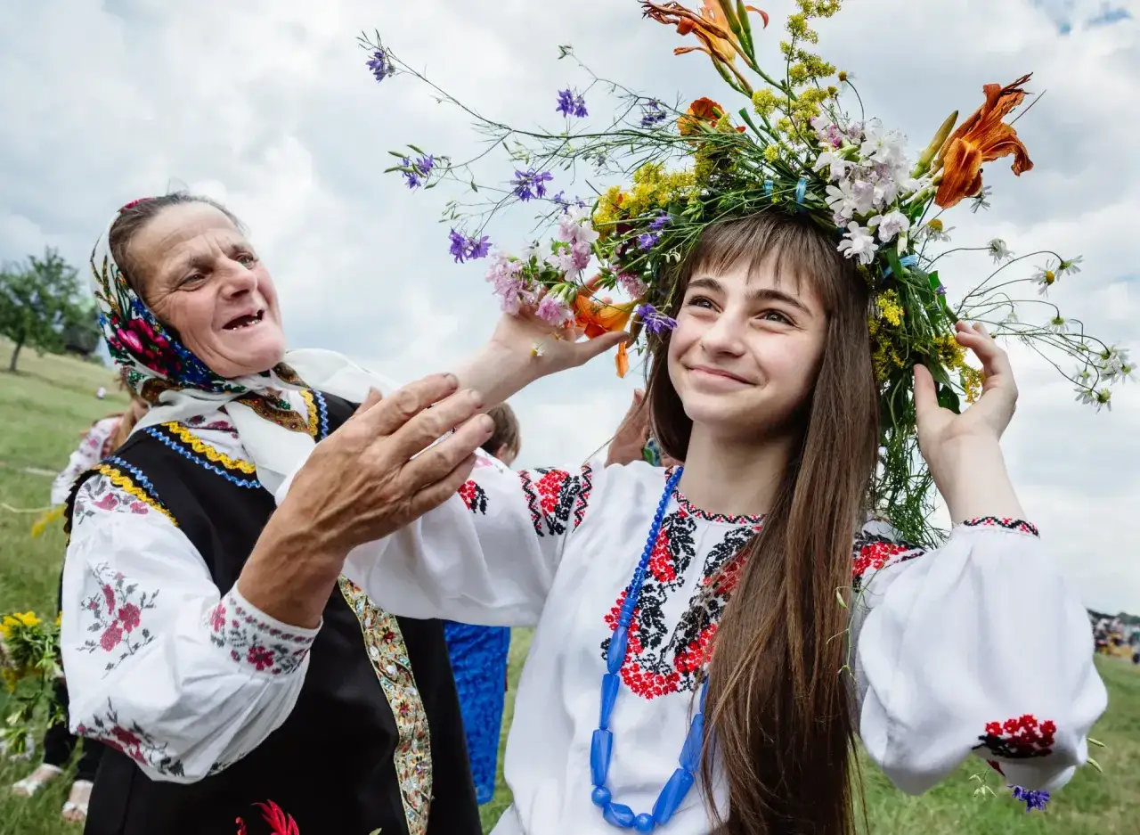 Starsza kobieta poprawia wianek z kwiatów młodej dziewczynie, gdy są sobótki. Obie ubrane w tradycyjne stroje.