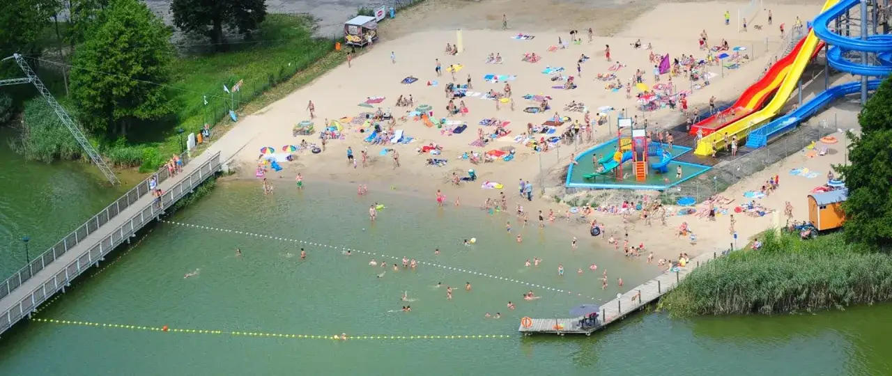 Tłum ludzi wypoczywa na piaszczystej plaży nad jeziorem Margonińskim. Widać zjeżdżalnie wodne, pomost i kąpielisko.