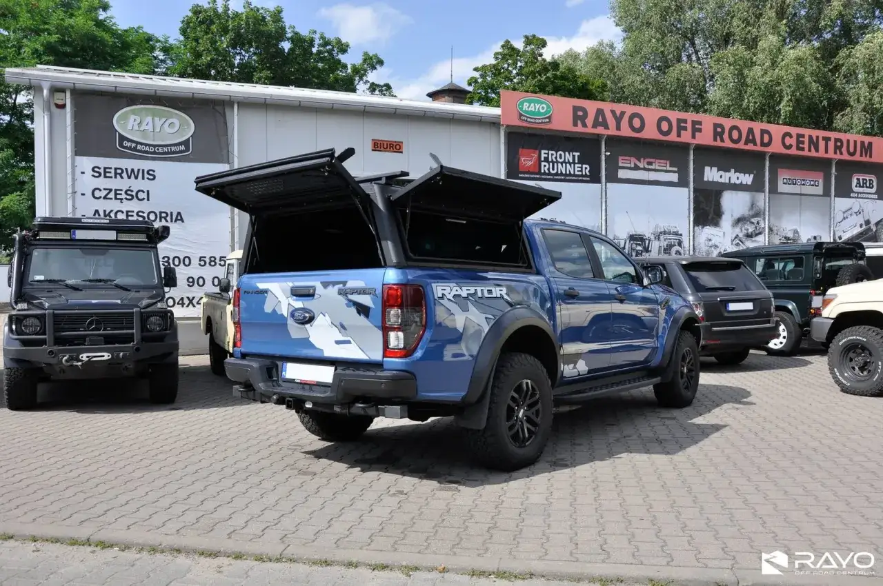 Niebieski Ford Ranger Raptor z otwartą klapą paki, zaparkowany przed centrum off-road. Obok czarny Mercedes G-klasa.