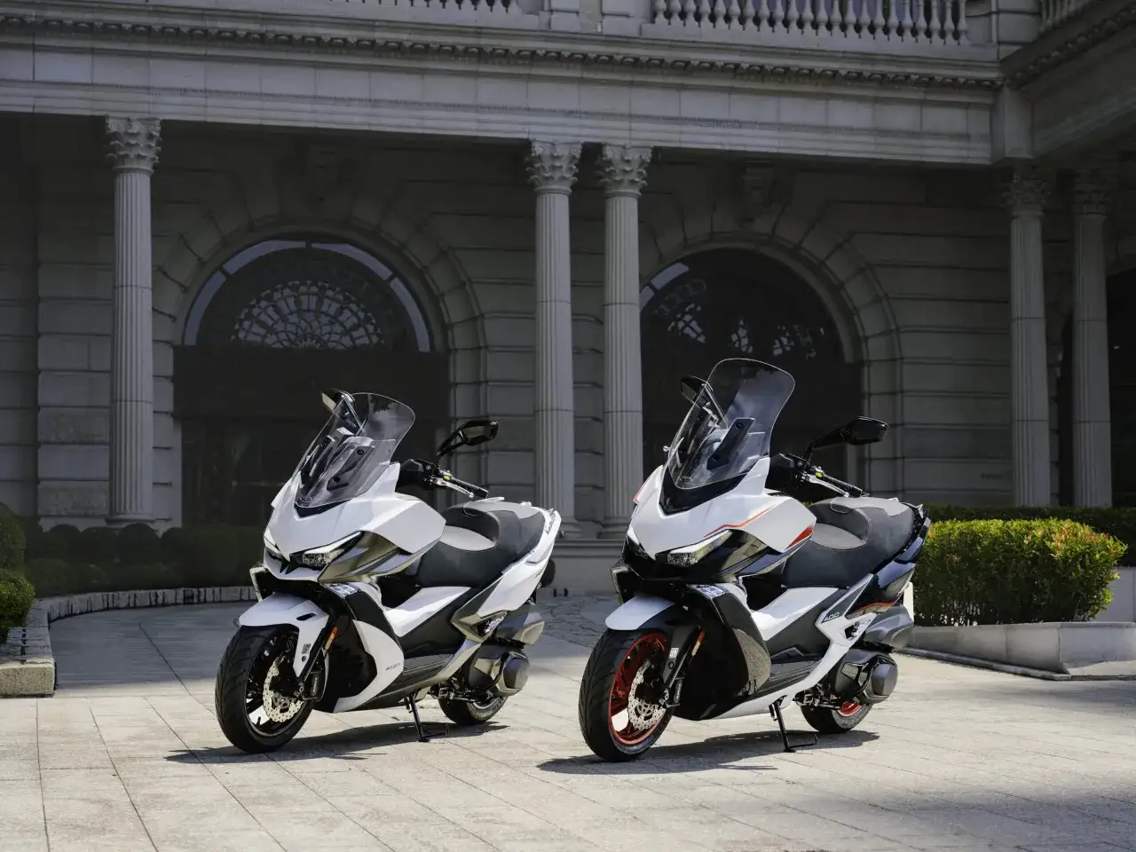 Dwa białe skutery Kymco Xciting 400i stoją przed eleganckim budynkiem. Poznaj kymco xciting 400i dane techniczne.