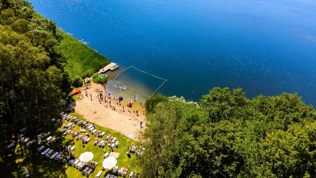 Słoneczna plaża i kąpielisko nad jeziorem, typowe dla mazurskich miejscowości. Ludzie odpoczywają na leżakach i kąpią się w wodzie.