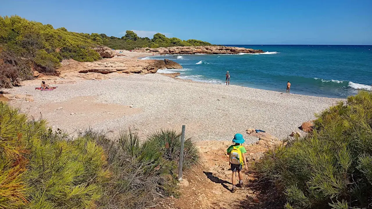 Niño con mochila azul desciende a la cala blanca de Castellón, mar turquesa y rocas.